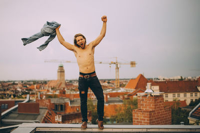 Full length of shirtless man holding shirt with arms raised standing on retaining wall at terrace