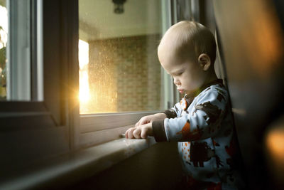 Baby boy with mobile phone standing by window at home