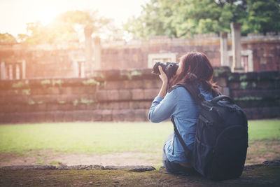 Rear view of female tourist photographing building through slr camera while sitting in forest
