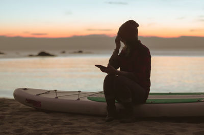 Woman sitting on shore at beach against sky during sunset