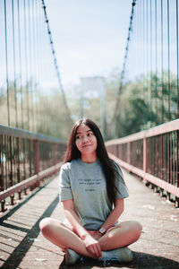 Portrait of smiling young woman sitting on footbridge against sky