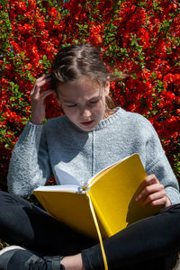 Young school age girl reading from yellow textbook in park against bright red flowers background.