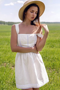 Young woman with long black hair stands in white dress and clothes straw hat is on the green field 