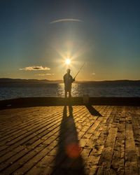 Silhouette man fishing at sea shore against sunset sky