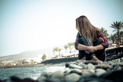 Woman sitting on rock at shore against clear sky