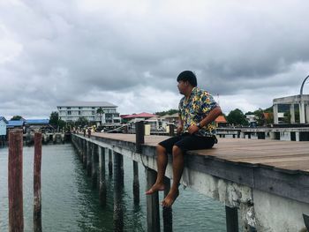 Man sitting on pier over sea against cloudy sky