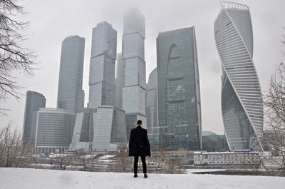 Rear view of man standing against skyscrapers during snowfall