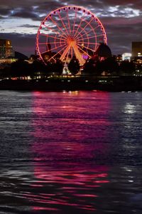 Illuminated ferris wheel by river against sky at night