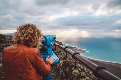 Rear view of woman looking through coin-operated binoculars standing by sea against sky