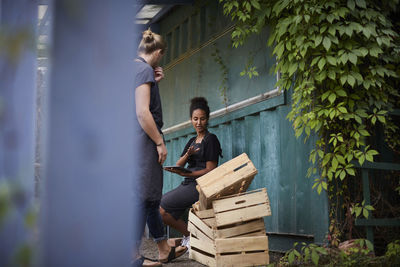 Female gardener holding digital tablet while communicating with friend over crates in yard