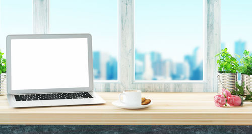 Close-up of laptop by coffee cup on table against window