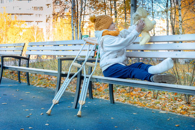 Kid walks in autumn park on crutches. child sitting on park bench. girl has one leg broken in cast.