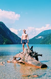 Portrait of shirtless young man standing on driftwood in lake against blue sky