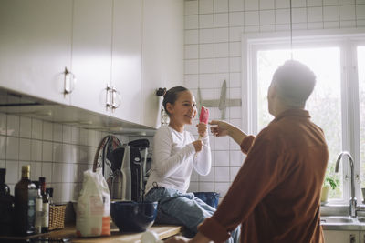 Smiling girl holding spatula while sitting on kitchen counter by father at home