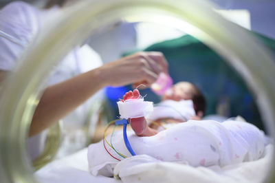 Unrecognizable medical worker feeding premature baby with tubes attached to foot in modern incubator while working in hospital