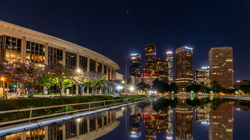 Illuminated modern buildings by river against sky at night