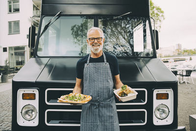 Portrait of smiling owner with food plate standing against commercial land vehicle