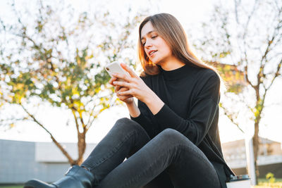 Portrait of young woman student with long hair using mobile phone in city park in golden hour