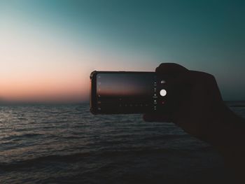 Cropped hand of person photographing sea against sky during sunset