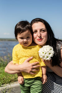 Portrait of smiling mother and daughter holding flower against sky