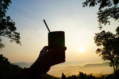 Silhouette person holding drink against sky at sunset