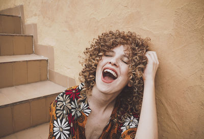 Cheerful young woman with curly hair and red lips sitting with eyes closed smiling in staircase against wall
