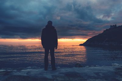 Rear view of man standing at beach against sky during sunset