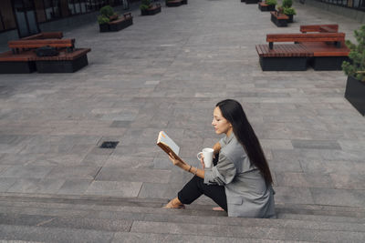 Businesswoman with coffee mug reading book on steps at office park