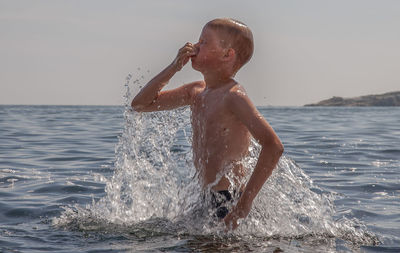 Full length of shirtless boy splashing water against sky