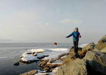 Boy throwing stone while standing on rocks at sea against sky