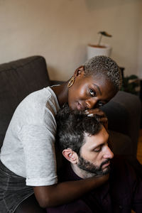 Young bearded man in casual shirt with closed eyes lying on comfortable couch in embrace with african american woman looking at camera in cozy living room