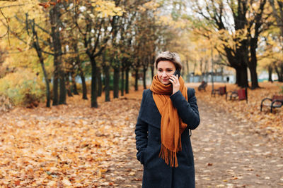 Short-haired blonde communicates on the phone walking in the autumn park in a coat and scarf