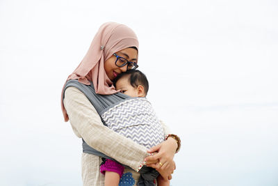 Mother wearing hijab carrying daughter against sky