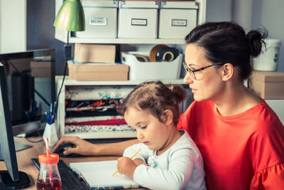 Mother teaching cute girl to write in book while sitting on table at home