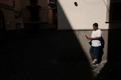 Full length of man using mobile phone while standing against wall