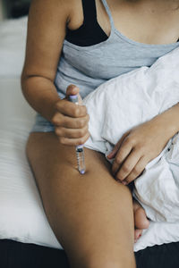 Midsection of woman injecting insulin in lap while sitting on bed in bedroom