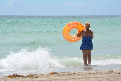 Rear view of woman with inflatable ring walking at beach against sky during sunny day