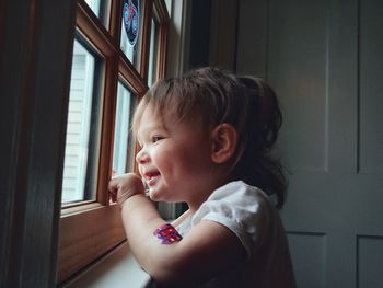 Side view of cute smiling baby girl looking through window at home