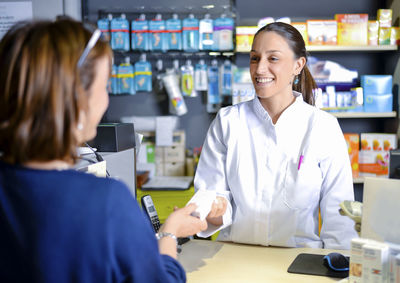 Portrait of a smiling female pharmacist standing while giving medicine to customer in store