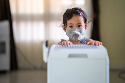 Cute little girl with healthy face mask standing breathing at air purifier. clean air concept.