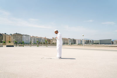 Full boy islamic male in traditional white clothes standing on rug and praying against blue sky on beach