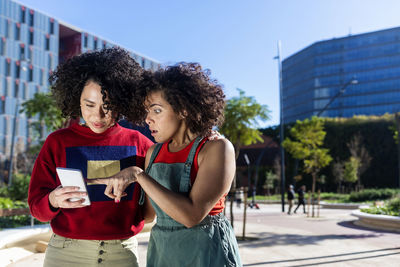 Smiling lesbian women looking at smart phone while standing against building