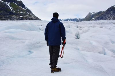 Rear view of man standing with axe at glacier against sky