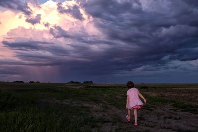 Rear view of girl walking on field against cloudy sky at dusk