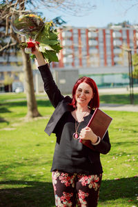 Portrait of smiling woman holding flower bouquet and book while standing in park