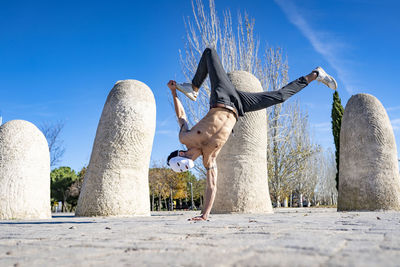 Shirtless man wearing white mask doing handstand while dancing on footpath during sunny day
