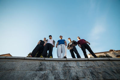 Low angle view of male friends standing on retaining wall against sky