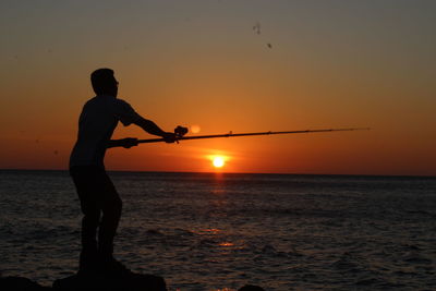 Silhouette man fishing in sea against sky during sunset