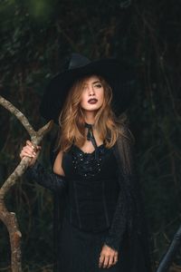 Young woman in hat looking away while holding tree trunk in forest