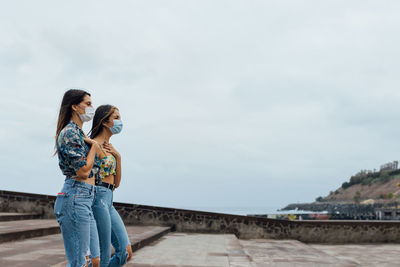 Side view of young slim girlfriends in trendy outfits and protective masks walking together on paved urban square during coronavirus outbreak